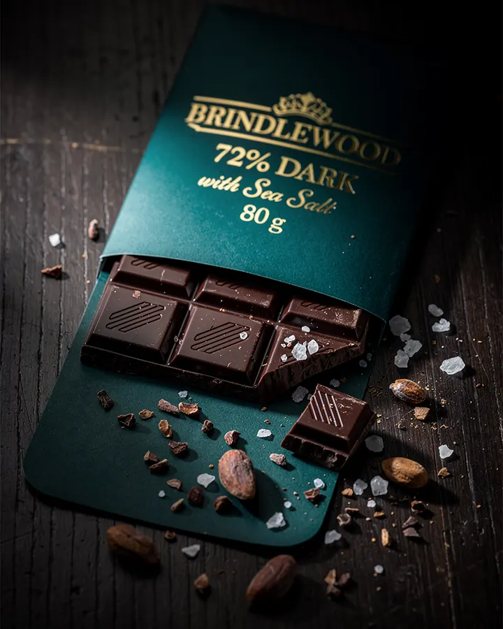 AI output for the third prompt: a teal wrapper with a gold crown drawn above the word Brindlewood and 72% Dark with Sea Salt 80 g in gold, with dark squares sliding out, salt and cacao nibs on dark wood, and no ad text.