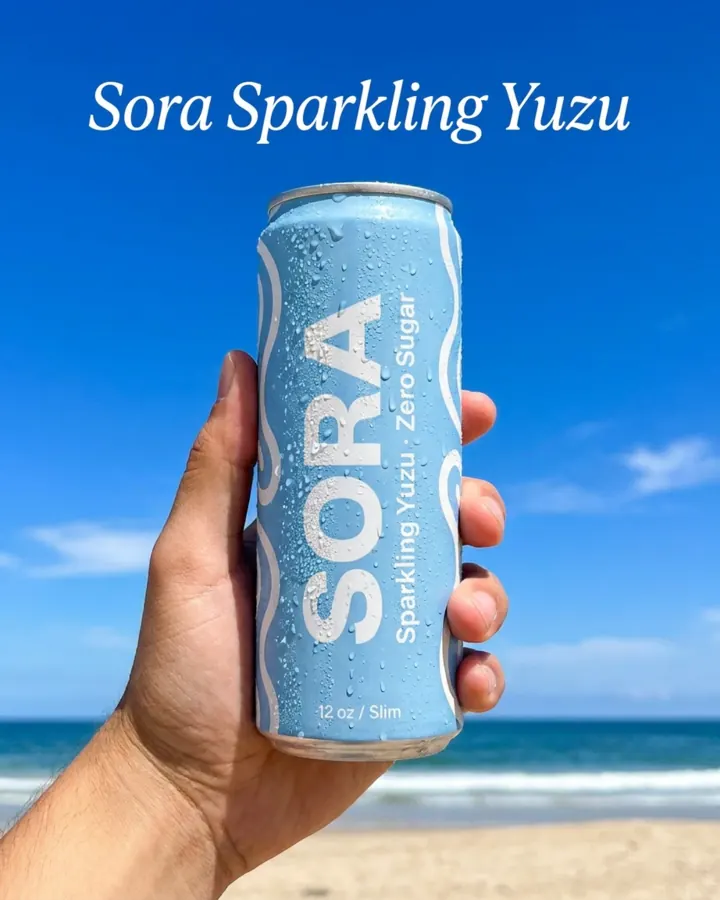 AI output for the fifth prompt: a hand holding Sora's light blue can, beaded with condensation, against a blue sky over a beach, under the line Sora Sparkling Yuzu.