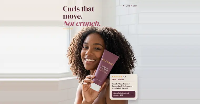 Soft white tiled bathroom look, Wildbraid ad: 'Curls that move. Not crunch.' A smiling woman with voluminous curly hair holds a plum Defining Curl Cream tube to her cheek. A card shows 4.7 stars, 2,641 reviews, and a 'Shop Defining Curl Cream, $26' button. The 1.91:1 version.